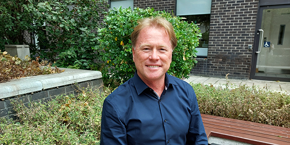 Barry Hingston is smiling in an outdoor courtyard while seated on a bench. Barry is wearing a dark navy shirt and there are trees and bushes surrounding him