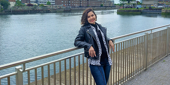 Camilla Eugenie standing and smiling while leaning on a metal rail along the Grand Canal Docks in Dublin. Camilla is wearing a leather jacket, a patterned blue and white shirt, and blue jeans