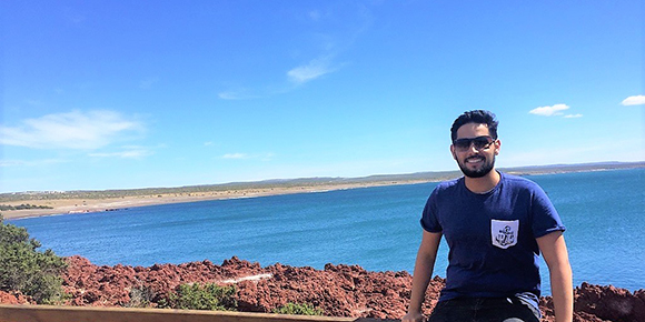 Guilherme Sobreira is smiling while sitting on a wooden ledge which has the background of the blue open sea behind him. He is wearing dark sunglasses and a dark blue t-shirt with a white pocket