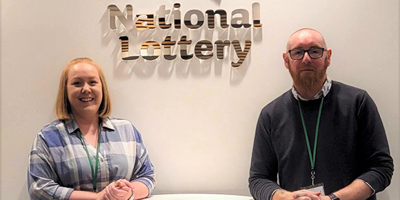 Helen Gaffney and Emmet Tannam are smiling beside each other with the National Lottery logo behind them. Helen is wearing a white and blue checked shirt and Emmet is wearing a black sweater with a white shirt collar popping out